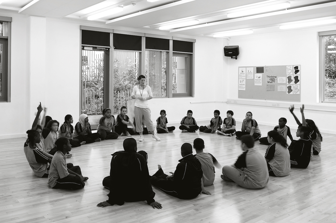 A group of secondary school students are sat in a circle on the floor with legs crossed, a few of them have their hands up. Their teacher is standing in the middle of the circle.